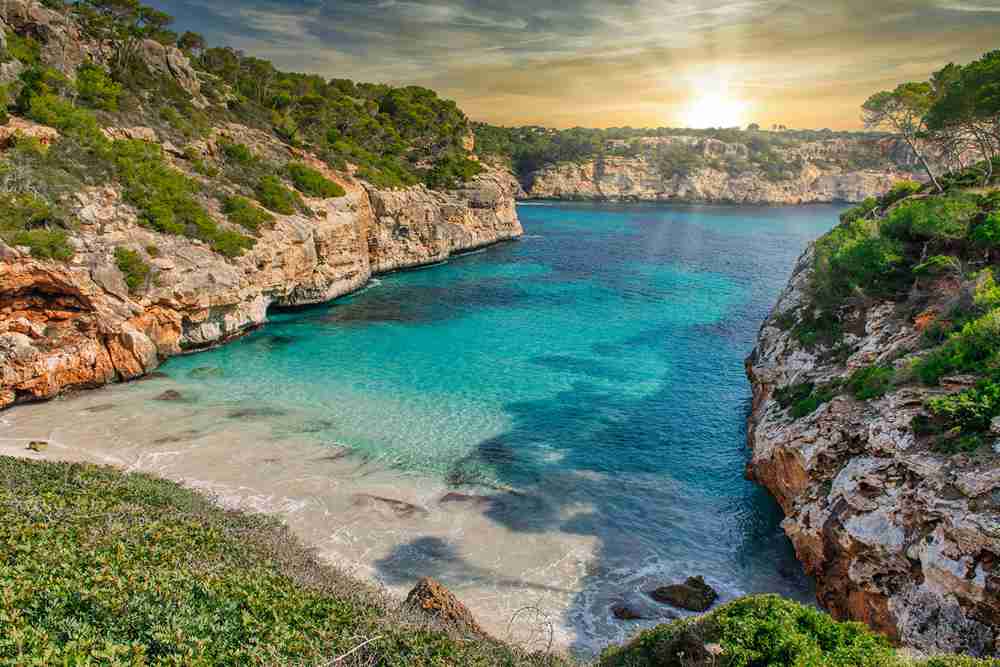 Immagine di un tramonto sulla spiaggia Paradise Calo des Moro a Maiorca, Spagna, con il mare e la costa rocciosa in primo piano.